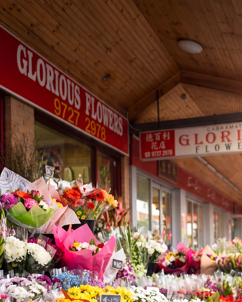 The red Glorious Flowers sign and the hanging sign under the awning, buckets of flowers on the footpath